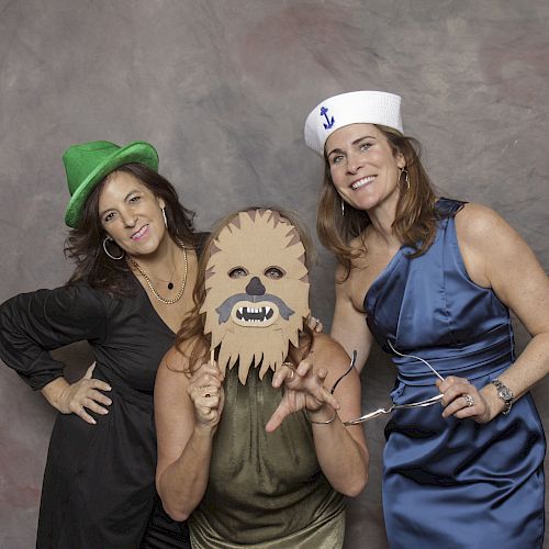 Three women pose for a fun photo, one in a green hat, one with a Chewbacca mask, and another in a sailor hat, against a plain backdrop.