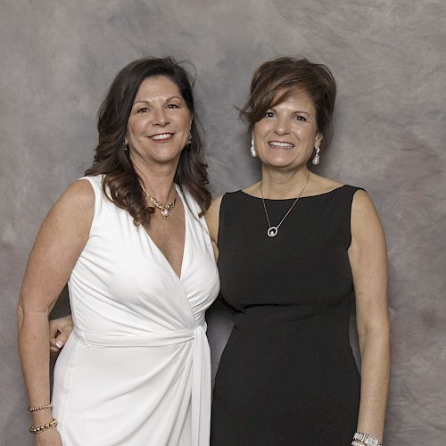 The image shows two women standing close together, smiling, one in a white dress and the other in a black dress, against a neutral background.