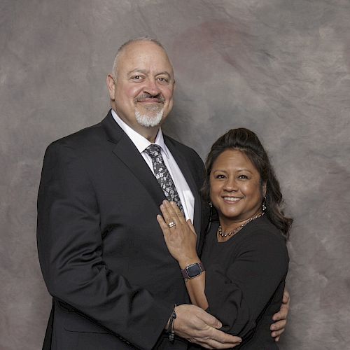 A man and a woman dressed in formal attire are standing together and smiling in front of a textured backdrop.