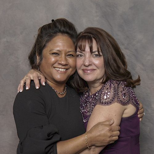 Two women are smiling and hugging each other in front of a gray backdrop.