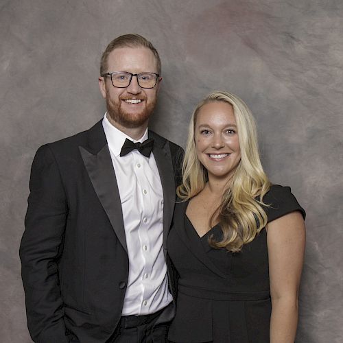 A man and woman are dressed formally and smiling for the camera against a gray textured backdrop.