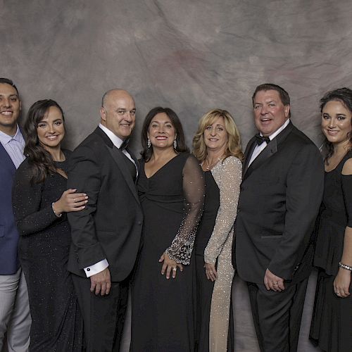 A group of seven people, dressed in formal attire, are posing together against a grey backdrop, smiling and standing close to each other.