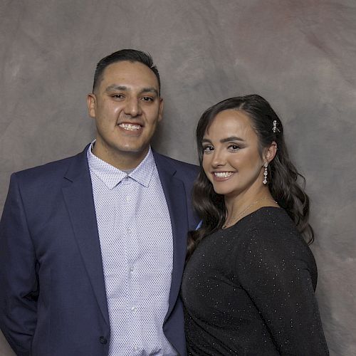 Two people are standing and smiling. The man is wearing a blue suit, and the woman is in a black dress.