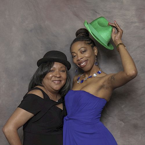 Two people posing together; one in a black dress and hat, the other in a blue dress holding a green hat.