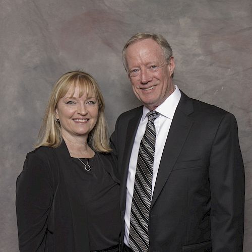 A smiling man and woman are standing side by side in formal attire, against a gray background.