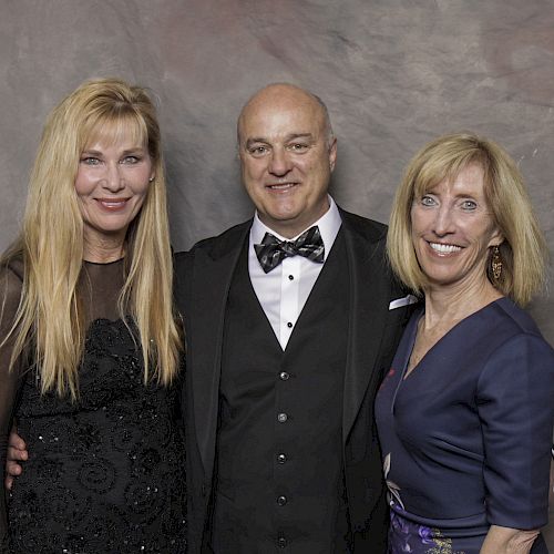 A man in a tuxedo and two women in formal dresses are posing together, smiling in front of a neutral backdrop, creating a formal group photo.