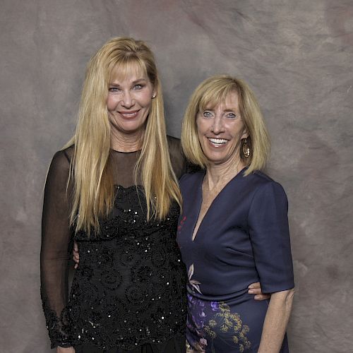 Two women are standing close together, smiling at the camera against a neutral background, one wearing a dark dress and the other a blue dress.