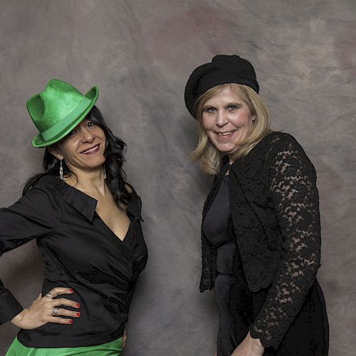 Two people are posing against a neutral backdrop. One wears a green hat and black outfit, while the other wears a black hat and dark clothing.