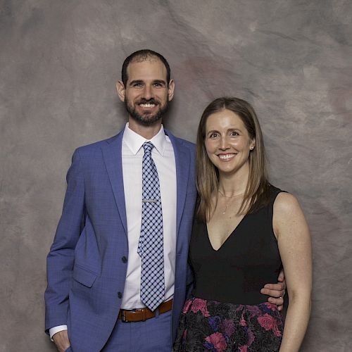 A smiling couple stands in front of a grayish backdrop, with the man in a blue suit and the woman in a black dress with a floral skirt.