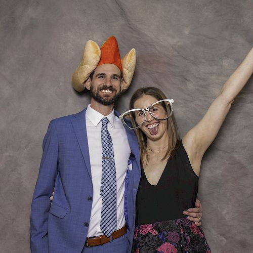 A man wearing a turkey hat and a woman wearing large novelty glasses pose for a fun photo; she points upwards with her tongue out, while he smiles.