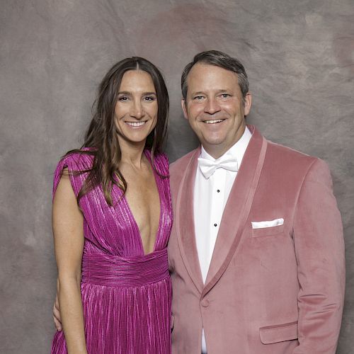 A woman in a pink dress stands next to a man in a pink suit with a bowtie, both smiling and posing for the photo.