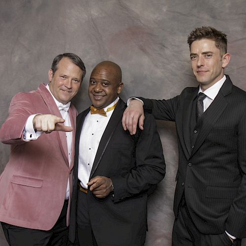 Three men in suits pose against a gray backdrop; one wears a pink jacket, one has a yellow bow tie, and the third is in a black suit.