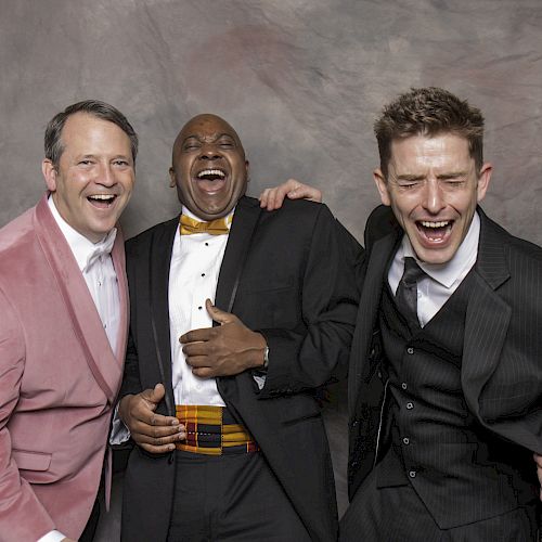 Three men in suits laughing together in front of a gray textured backdrop. They appear to be enjoying themselves, sharing a happy moment.