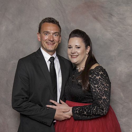 A man in a black suit and tie stands beside a woman in a black lace top with a red skirt, both smiling against a neutral backdrop.