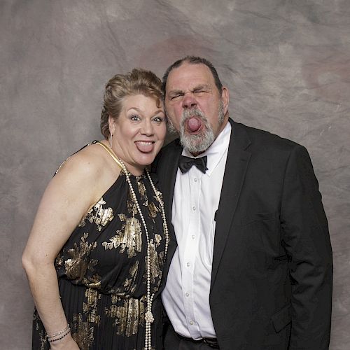 Two people are dressed formally, both making funny faces with their tongues sticking out, in front of a neutral backdrop.