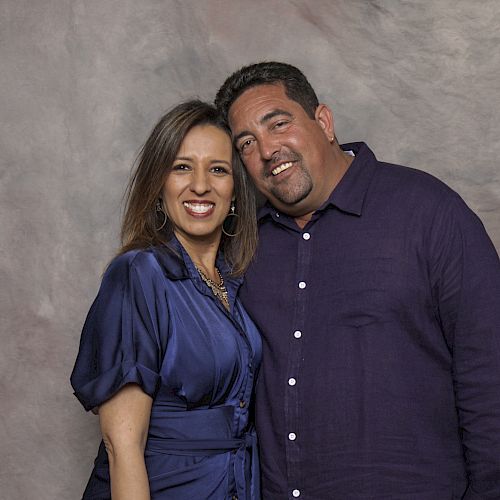 In this image, a man and a woman are standing close together, smiling at the camera, with a neutral-colored background.