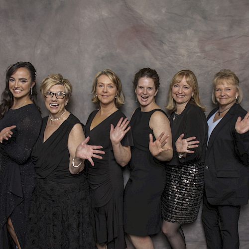 A group of six women dressed in formal attire are standing in front of a gray backdrop, smiling and waving at the camera.