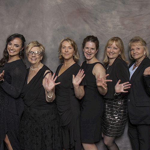 Six women in formal attire, standing in a line, smiling, and holding their hands up as if waving or gesturing, with a neutral backdrop behind them.