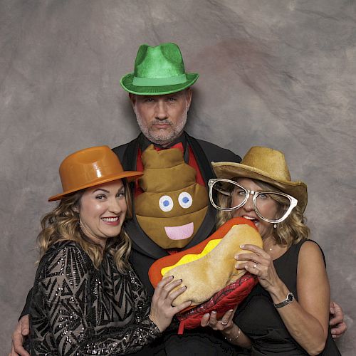 Three people posing with props: green, orange, and yellow hats, large glasses, plush hot dog, and a poop emoji. They are smiling and playful.