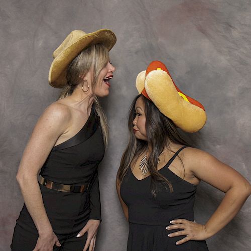 Two women in black dresses, one wearing a cowboy hat and the other a hotdog hat, posing against a grey backdrop, one laughing.