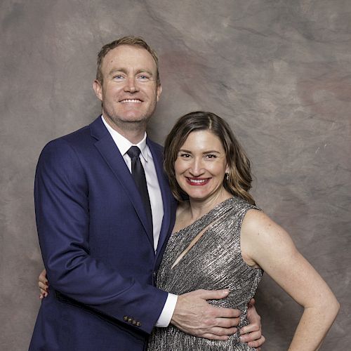 A smiling couple poses together in formal attire against a neutral backdrop.