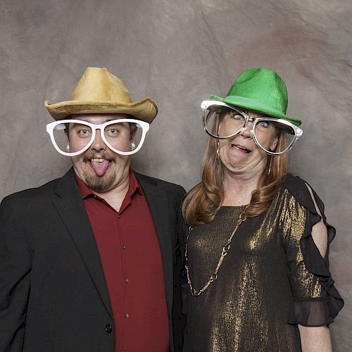 Two people wearing oversized glasses and hats, making funny faces, dressed formally, posing against a plain backdrop.