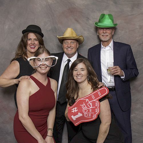 Five people are posing for a photo booth image; some are wearing hats and novelty glasses, and one has a #1 foam finger.