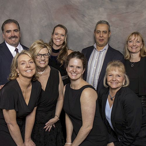 A group of eight people, dressed formally, posing together in front of a greyish backdrop. Everyone is smiling and appears to be in good spirits.