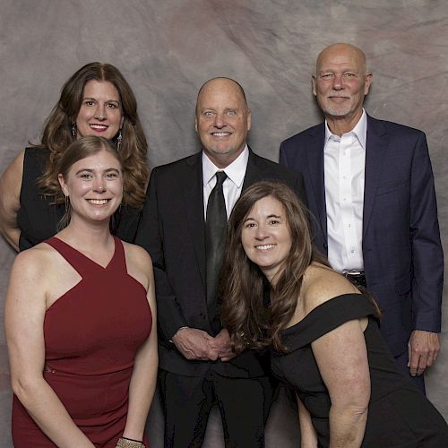 A group of five people, three women and two men, dressed in formal attire and posing for a photo against a gray backdrop.