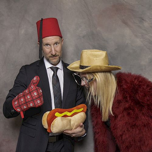 Two people in costumes: one in a suit with a fez and holding a hot dog plush, the other in a hat and fur coat, giving a thumbs-up.