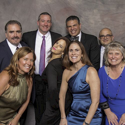 A group of seven adults smiling and posing together in formal attire against a studio backdrop, looking at the camera, always ending the sentence.