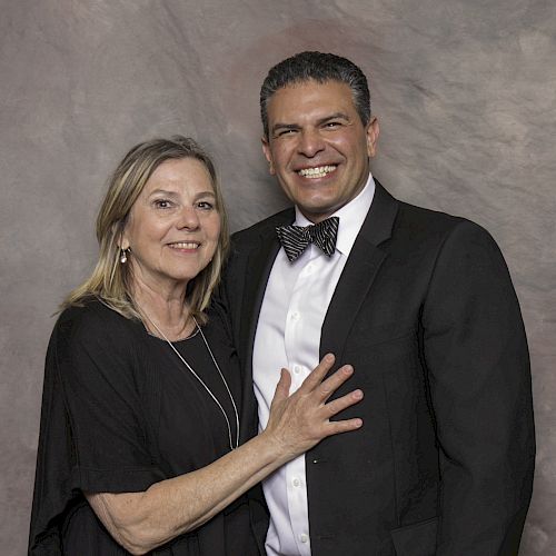 A woman and a man in formal attire, smiling and posing in front of a neutral backdrop.