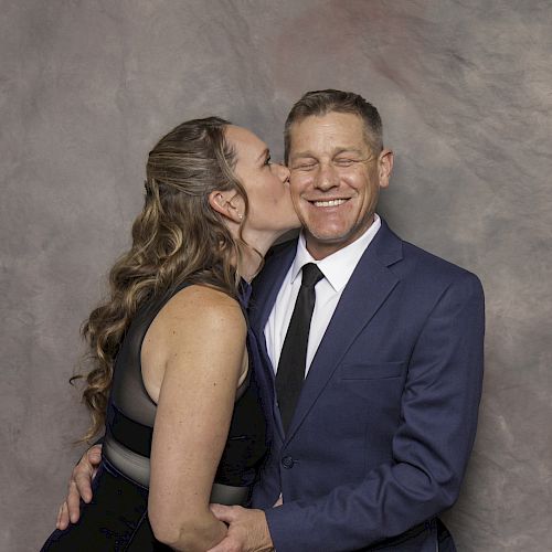 A woman in a black dress is kissing a man in a blue suit and tie on the cheek while he smiles.