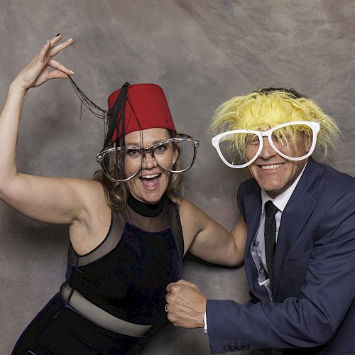 Two people dressed playfully with oversized glasses and wigs, posing enthusiastically against a neutral backdrop.