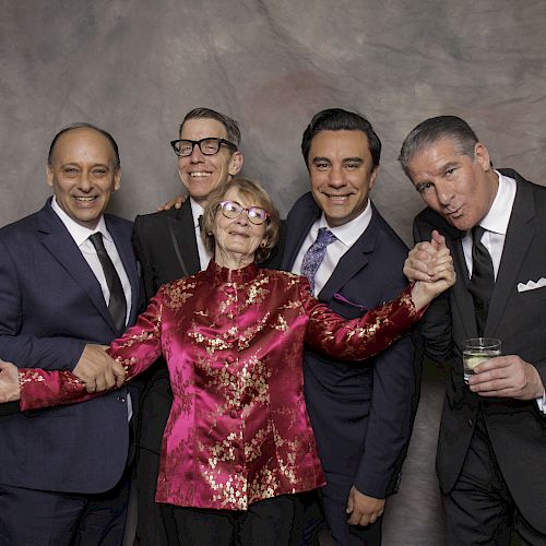 A group of five people, four men in suits and one woman in a red blouse, pose together for a photo in front of a gray backdrop, smiling and cheerful.