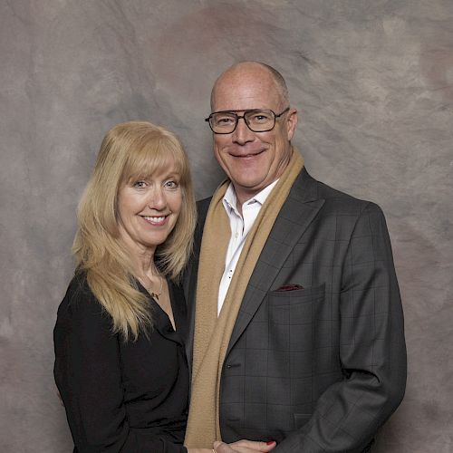 A couple smiling at the camera, standing against a neutral backdrop; the man wears a suit and scarf, and the woman wears a black outfit.