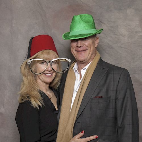 Two people pose for a photo with silly hats and exaggerated glasses, smiling against a neutral backdrop.