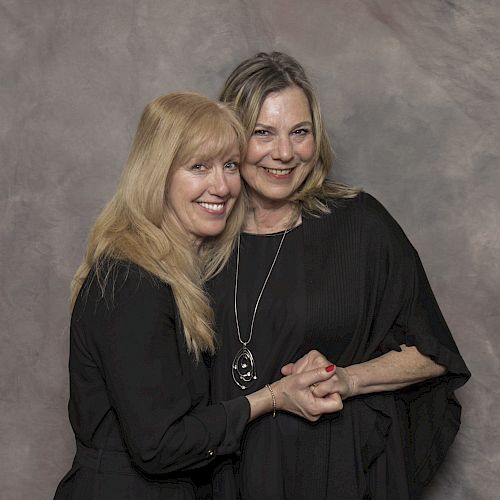 Two women are posing together, smiling and holding hands, against a neutral backdrop.