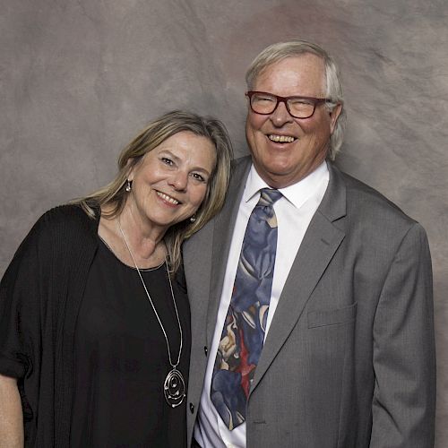 A smiling man in a suit and woman in a black dress are posing together against a gray textured background.