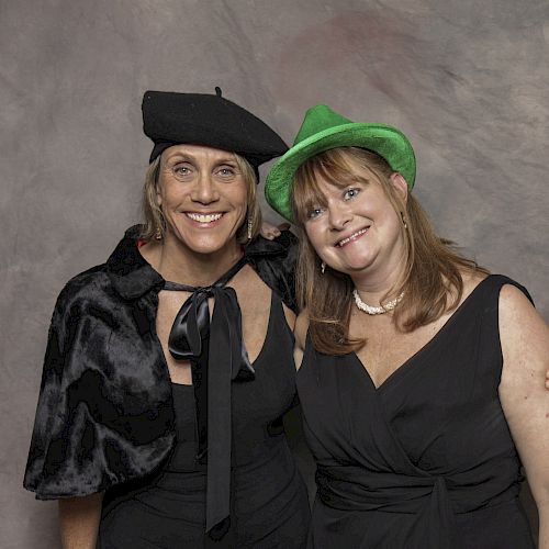 Two women in black outfits, one wearing a black beret and the other in a green fedora, smiling and posing against a neutral backdrop.