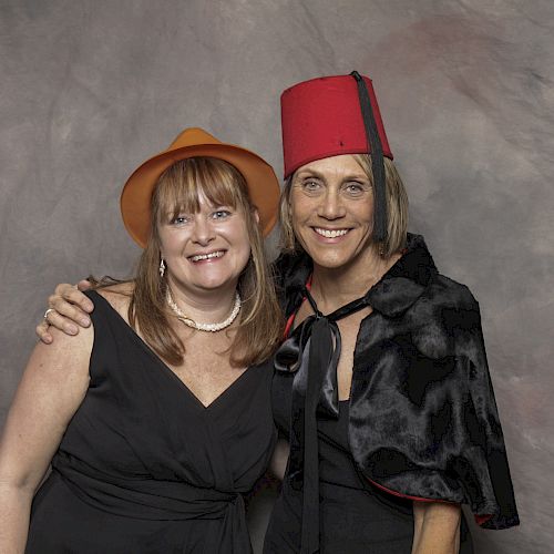 Two women are posing together, smiling at the camera. One is wearing an orange hat and the other a red fez and a cape, in front of a gray backdrop.