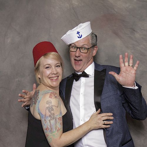 Two people are posing together, wearing hats and formal attire, with the man making a playful gesture and the woman smiling.