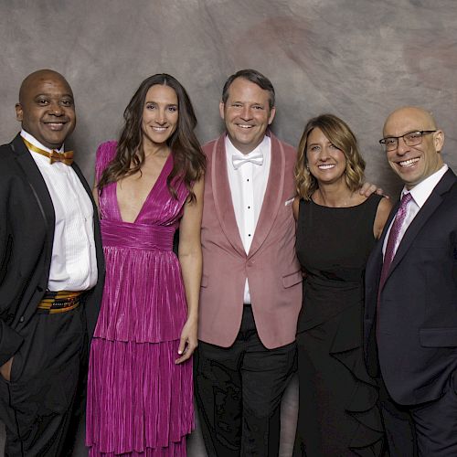 Five people dressed in formal attire, posing against a gray backdrop, all smiling at the camera.