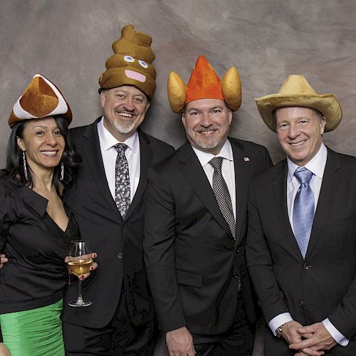 Four people dressed formally, wearing novelty hats such as a poop emoji and turkey hat, smiling at the camera.