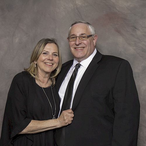 A smiling man in a suit and a woman in a black dress pose for a portrait in front of a grayish-brown background, looking pleasantly at the camera.