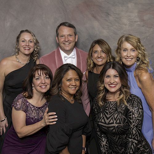 Seven people are posing together in formal attire against a backdrop, smiling at the camera.