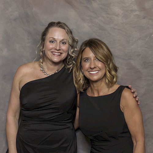 Two women are standing together, smiling, and posing for a photo in front of a grayish background.